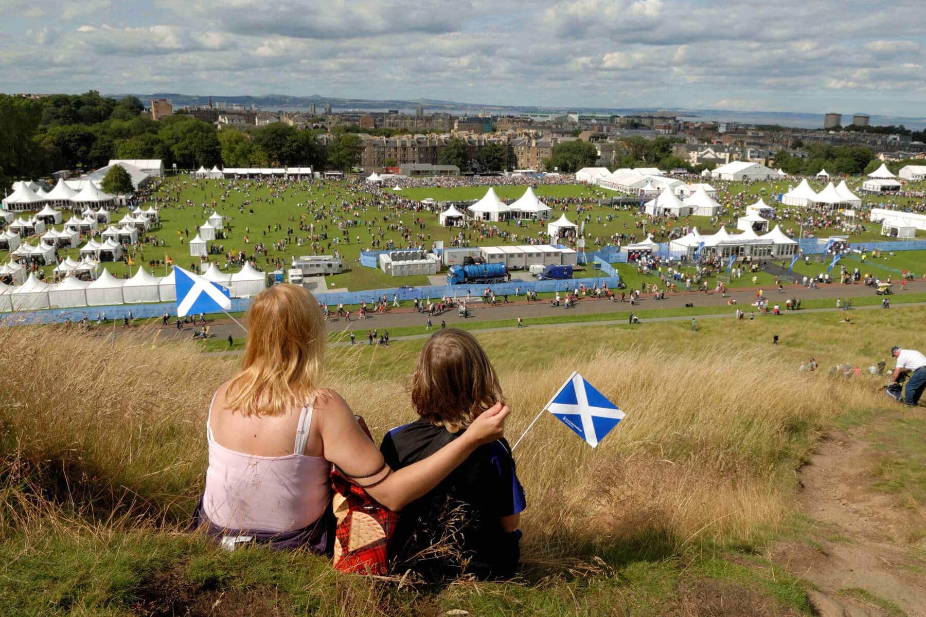 A photo of an woman and child holding small Scottish saltire flags, sitting on a hillside watching the crowds and tents of The Gathering 2009