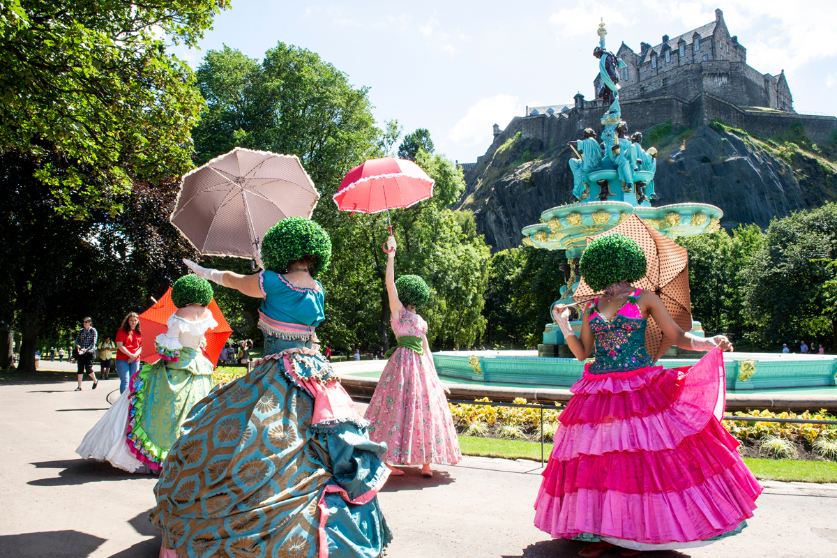 A photo of four dancers, wearing extravagant dresses and face obscuring helmets which look like bushes, in Princes Street Gardens, Edinburgh, at the launch of the newly renovated Ross Fountain