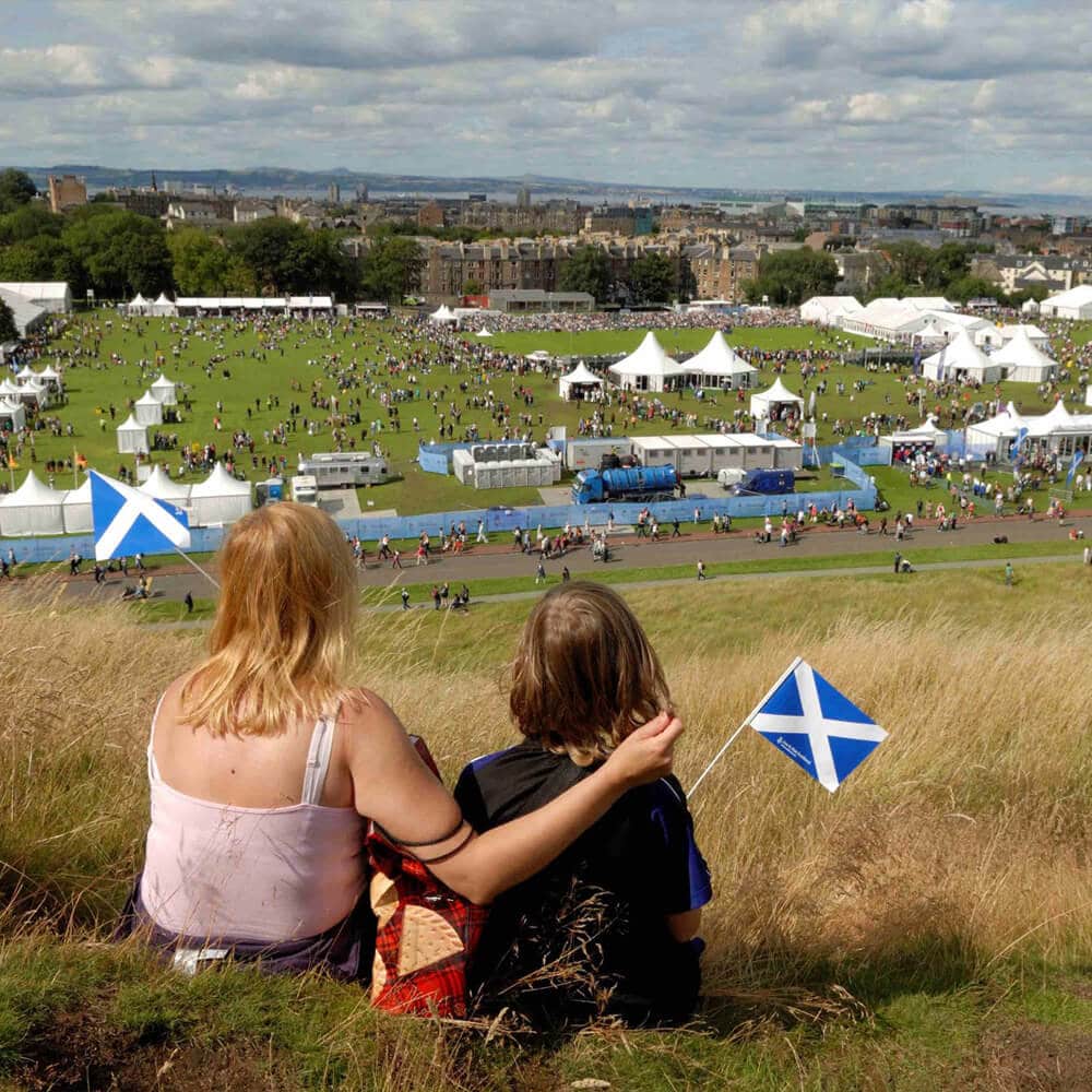A photo of an woman and child holding small Scottish saltire flags, sitting on a hillside watching the crowds and tents of The Gathering 2009